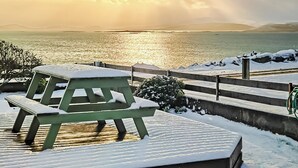 Una mesa y banco de picnic cubiertos de nieve junto a la orilla, con un atardecer.