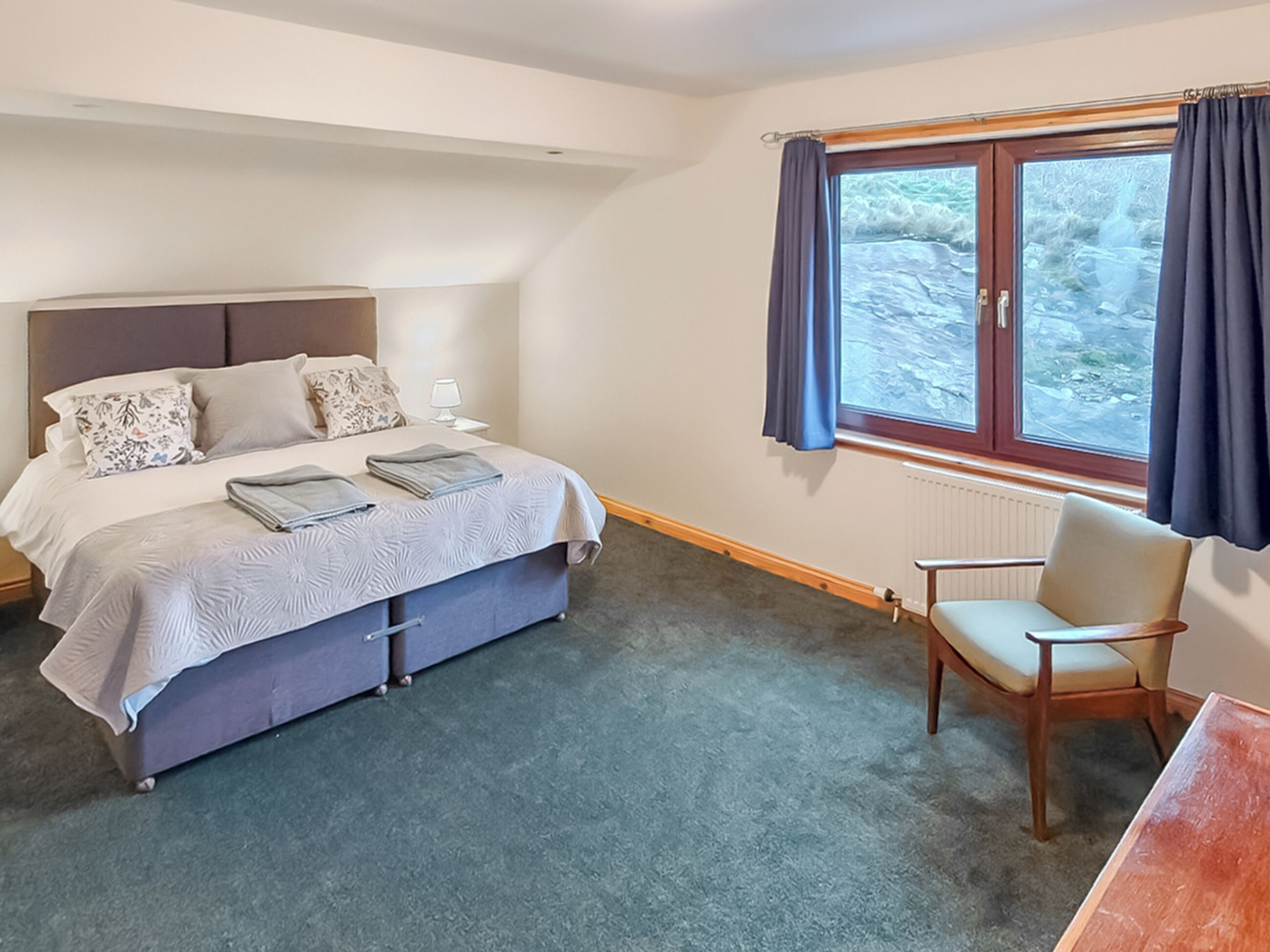 Une chambre à coucher avec un lit, une table de chevet, une lampe, une chaise et une fenêtre avec des rideaux.