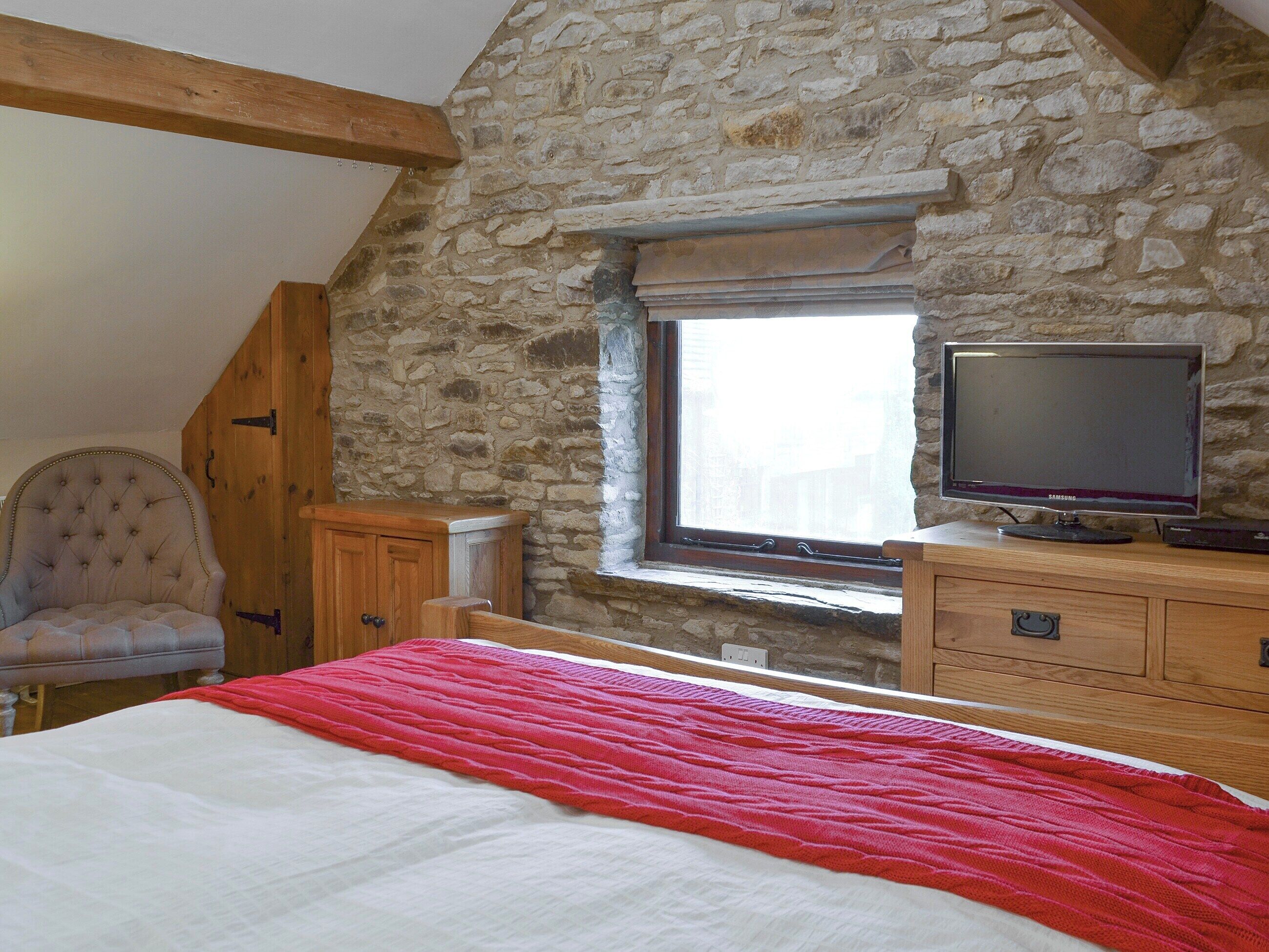 A bedroom with a stone wall, a bed with a red blanket, a wooden dresser, a television, and a chair.