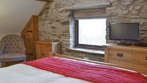 A bedroom with a stone wall, a bed with a red blanket, a wooden dresser, a television, and a chair.