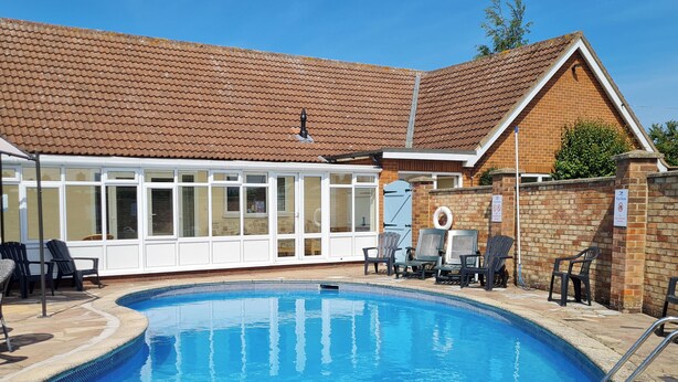 Ein Swimmingpool mit Fliesen umgeben, mit Liegestühlen und einer überdachten Terrasse mit Glasüren.