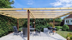 Eine überdachte Terrasse mit Gartenmöbeln, einem Grill und einer Pergola.