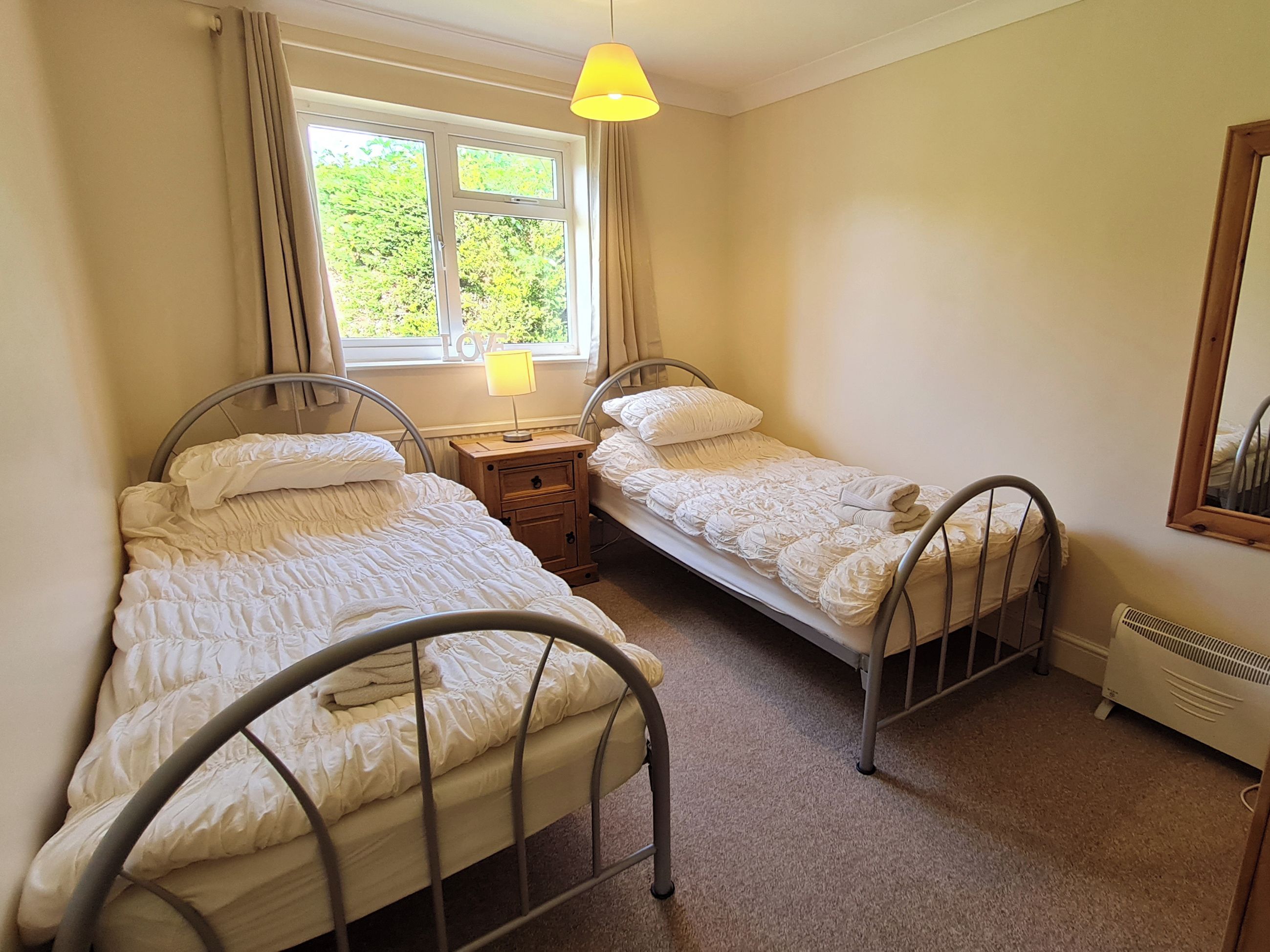 A room with two metal-framed beds, a small wooden nightstand, a window with curtains, and a radiator.