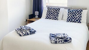Ein ordentlich bezogenes Bett mit gemusterten Handtüchern, ein Nachttisch mit Lampe und eine herzförmige Dekoration an der Wand.