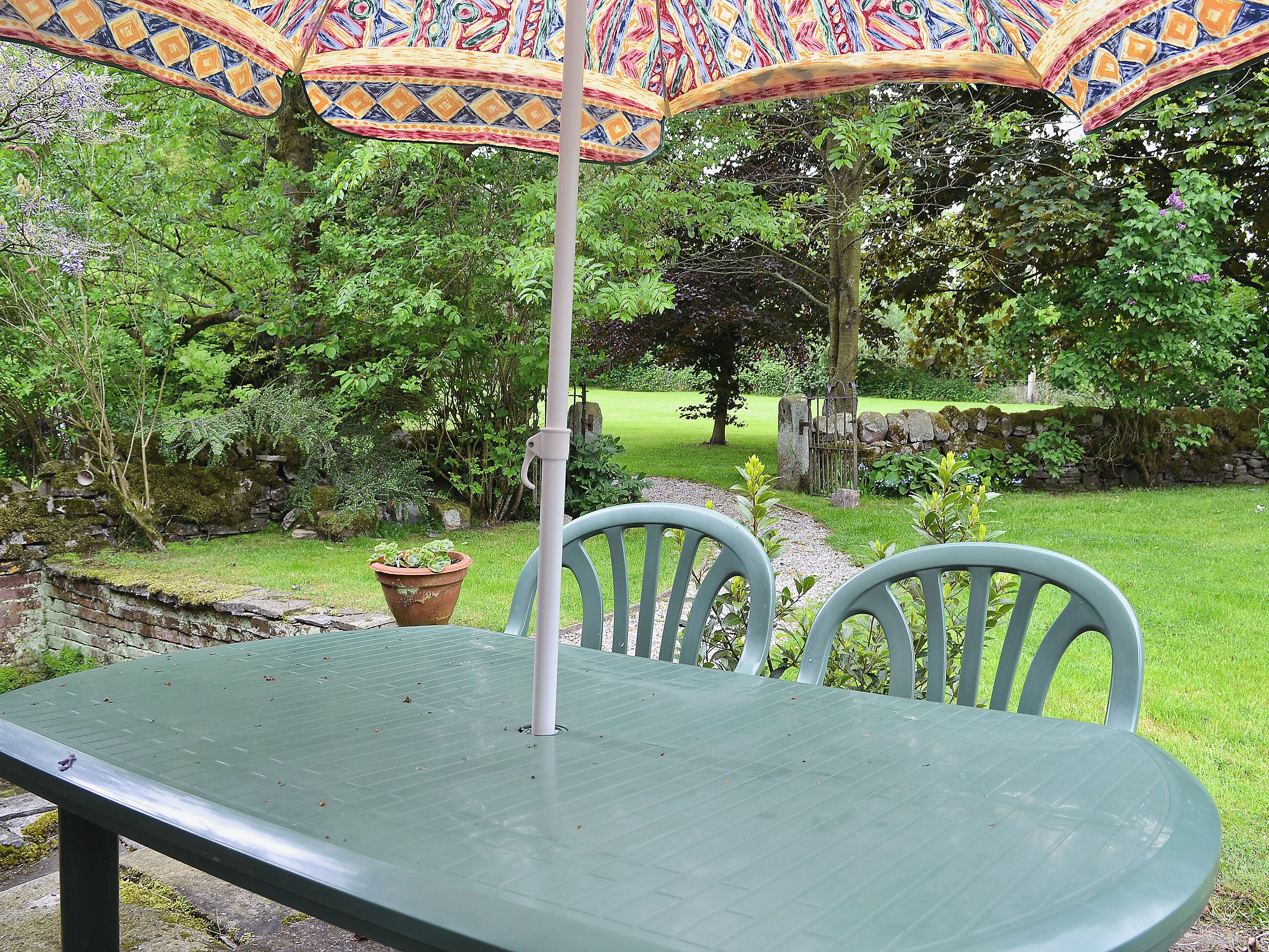 An outdoor patio with a green table and two chairs, an umbrella, and a stone wall in the background.