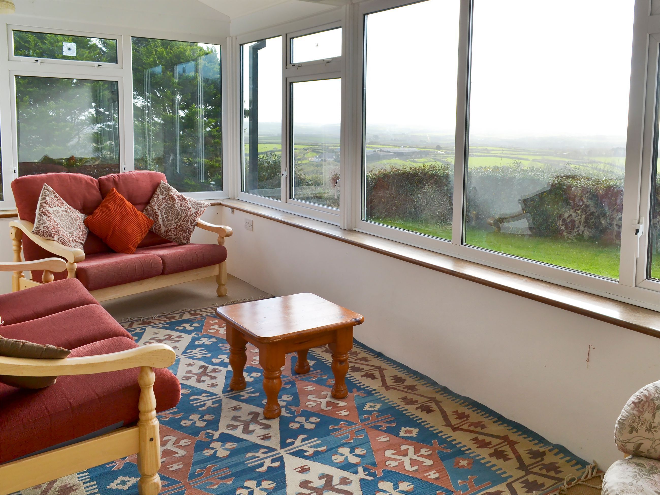 Ein Zimmer mit einem roten Sofa, einem Holzstuhl und einem kleinen Tisch. Große Fenster bieten Blick auf eine grüne Landschaft.