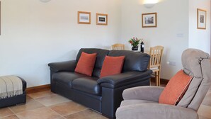 Ein Wohnzimmer mit einem schwarzen Ledersofa, einem grauen Sessel und einem Holzesstisch mit Stühlen. An den Wänden hängen Bilder in Rahmen und auf dem Tisch steht eine Vase mit Blumen.