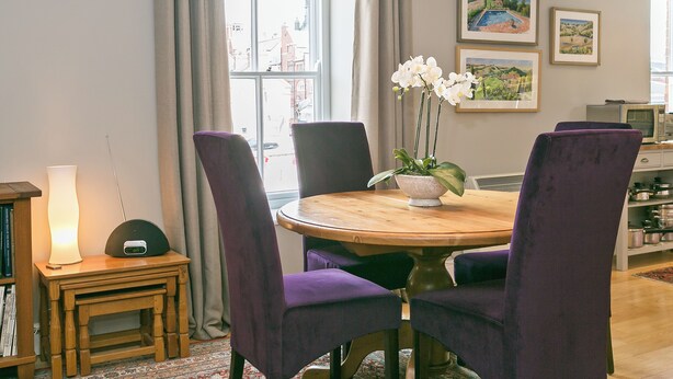 A dining area with a round wooden table, purple upholstered chairs, a small side table with a lamp, and a vase with white flowers. - Jorvik- Woodsmill Quay (Skeldergate, York)