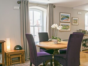 A dining area with a round wooden table, purple upholstered chairs, a small side table with a lamp, and a vase with white flowers.