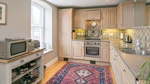 A kitchen with light wooden cabinets, a microwave, oven, and a sink. There is a patterned rug on the floor and a window providing natural light. - Jorvik- Woodsmill Quay (Skeldergate, York)