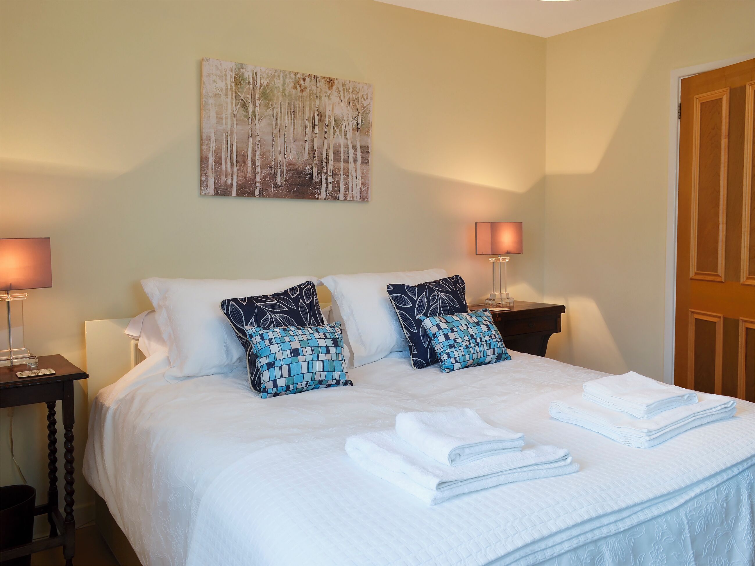 A neatly made bed with white linens and two patterned pillows, flanked by bedside tables with lamps, in a room with a large painting on the wall.