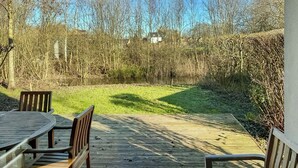 Una terraza de madera con una mesa y sillas, rodeada de árboles y un espacio cubierto de césped.