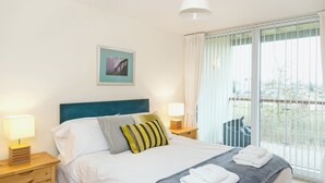 Una cama bien hecha con almohadas, una lámpara de noche, un cuadro en la pared y una ventana con cortinas.