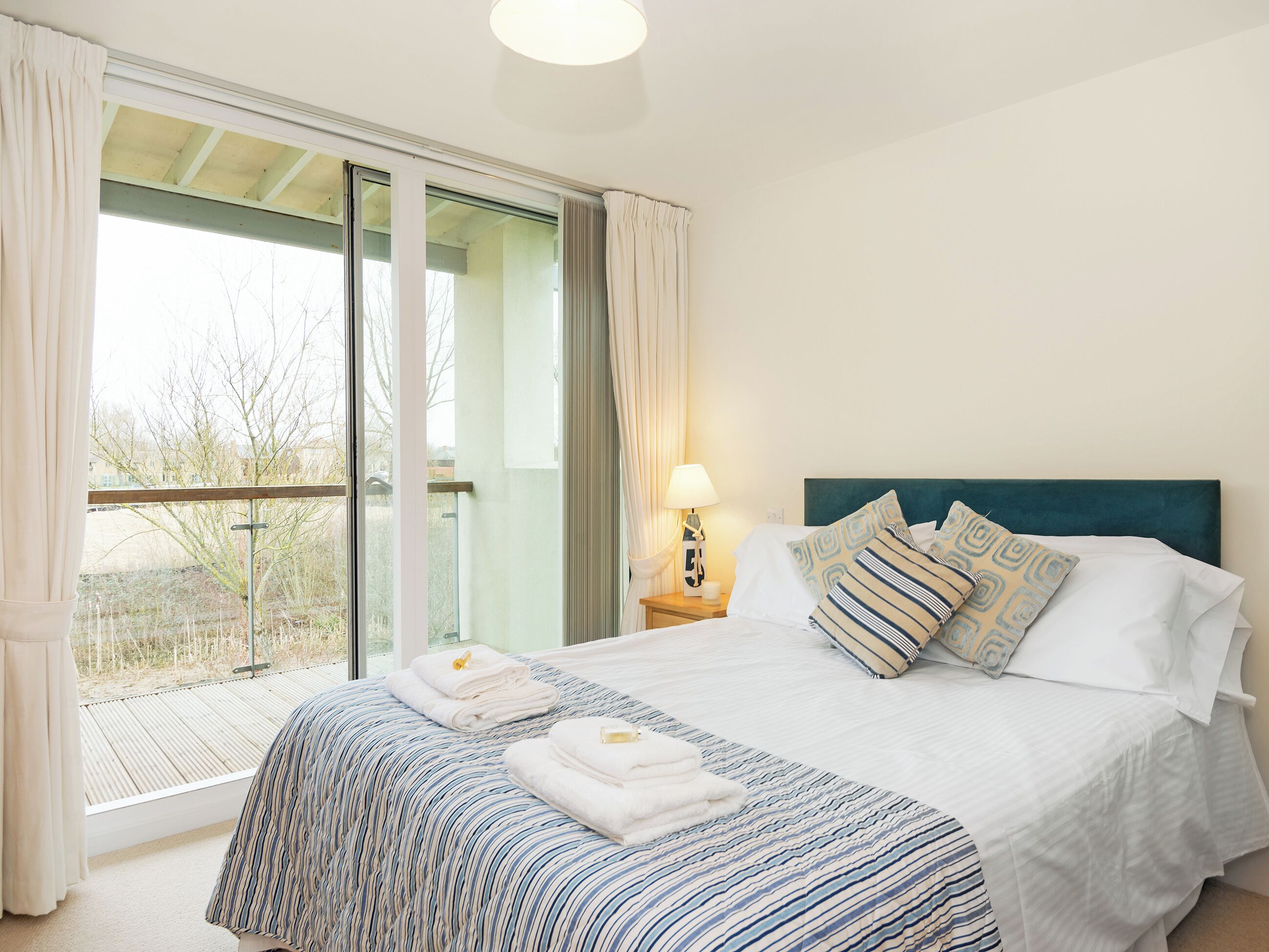 A hotel room with a large window, a bed with striped bedding, a nightstand with a lamp, and a view of the outdoors.