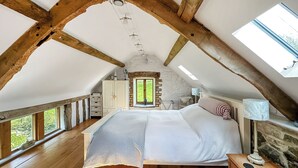 Un dormitorio con techo inclinado, vigas de madera, cama, cómoda, lámpara y ventana con vistas a la vegetación.