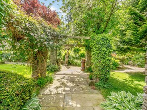 Ein gepflasterter Weg führt durch einen üppigen Garten mit einem hölzernen Torbogen und einer Statue.