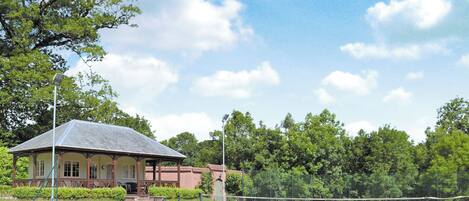 A tennis court with a pavilion, surrounded by trees and a clear sky.
