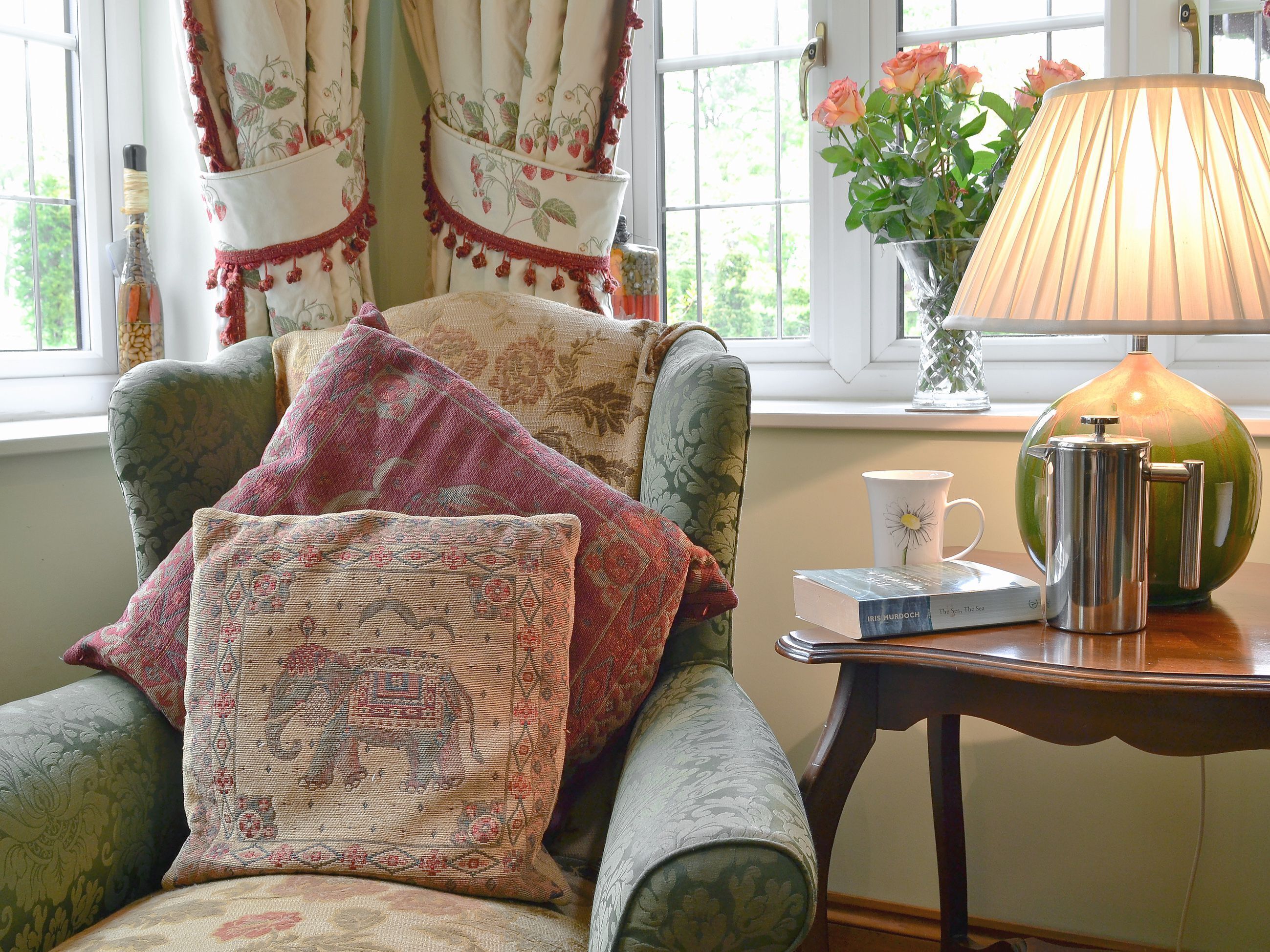 A cozy room with a green armchair, a lamp, a coffee pot, and a vase with flowers.
