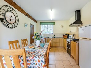 A kitchen with wooden cabinets, a white refrigerator, a dining table with a floral tablecloth, and a wall clock. - Granary Cottage - E5634 (Chittlehampton, near Umberleigh)