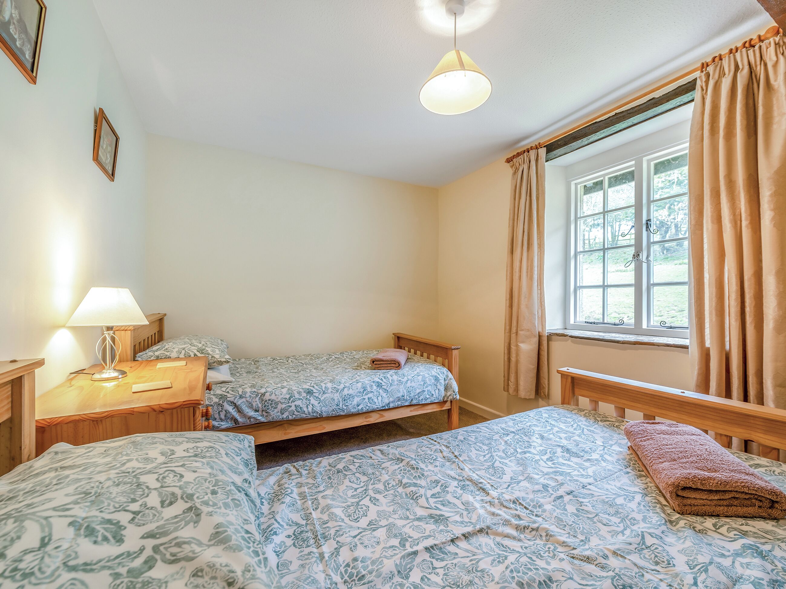 A bedroom with two beds, a bedside table with a lamp, a window with curtains, and a view of greenery outside.