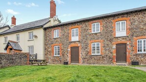 A stone building with multiple windows and doors, a grassy lawn in front, and a neighboring building with a tiled roof. - Granary Cottage - E5634 (Chittlehampton, near Umberleigh)
