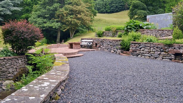 Un camino de grava que conduce a una zona con bancos de piedra, rodeada de abundante vegetación y un muro de piedra.