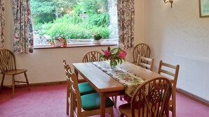 Comedor con mesa de madera, sillas con cojines verdes, cortinas con motivos florales y vista a zonas verdes.