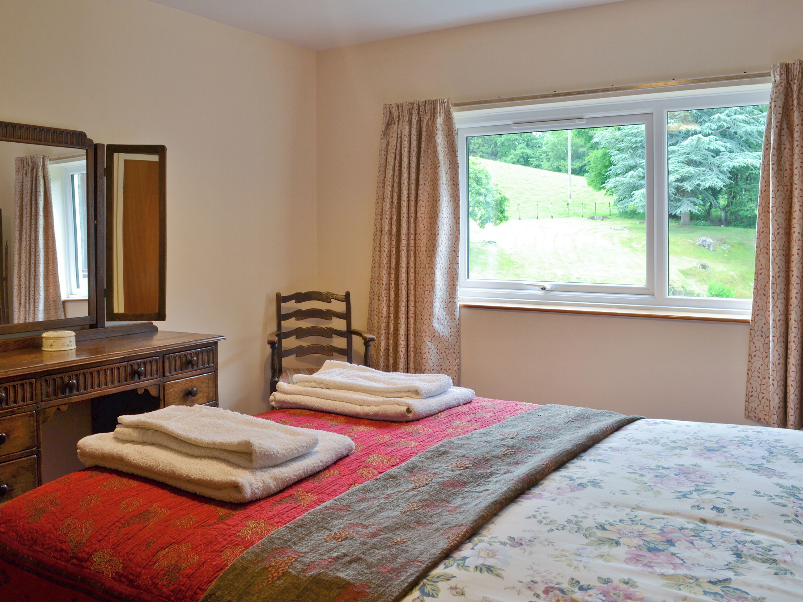 Une chambre à coucher avec un lit, une commode en bois, une chaise et une fenêtre donnant sur un paysage verdoyant.