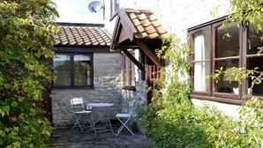 Ein Steinhaus mit Ziegeldach, eine kleine Terrasse mit zwei Stühlen und einem Tisch sowie ein Fenster mit Vorhängen.