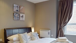 Un lit bien fait, avec du linge de lit blanc et un plaid en fourrure, une table de chevet avec une lampe, et un mur orné de trois œuvres d’art encadrées.