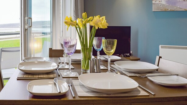 Une table à manger dressée avec des assiettes, des couverts et des verres, agrémentée d’un vase de fleurs jaunes et d’un tableau accroché au mur.