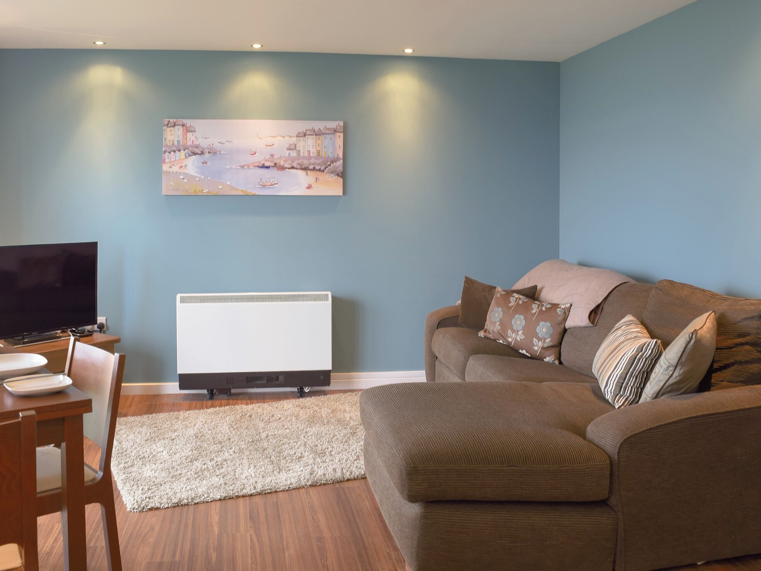Uma sala de estar com parede azul, uma TV de tela plana, um sofá marrom com almofadas estampadas, um radiador branco e uma mesa de jantar de madeira com cadeiras.