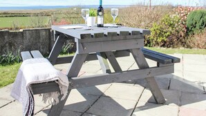 A wooden picnic table with a bottle and glasses on it, set on a patio with a view of the countryside.