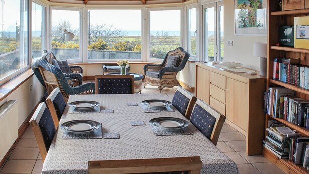 A dining area with a table set for four, surrounded by wicker chairs, a bookshelf, and a view of the outdoors.