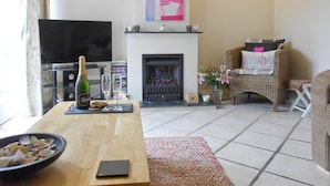 Ein Wohnzimmer mit Kamin, Fernseher, einem Esstisch mit einer Champagnerflasche und einem Korbstuhl.