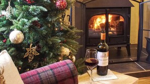 Un salon chaleureux avec un canapé à carreaux, un sapin de Noël, une cheminée et une bouteille de vin posée sur une petite table en bois.