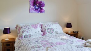 Ein Schlafzimmer mit blumigem Bettüberwurf, zwei Nachttischlampen, einem Nachttisch mit Schubladen und einem Wandbild mit Blumen.