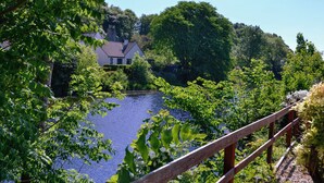 Blick auf einen Fluss mit einer Holzbrücke, üppigem Grün und einem Haus im Hintergrund.