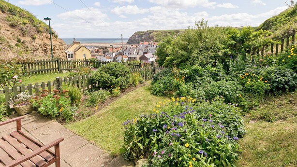 A garden with a wooden bench, a variety of flowers, and a view of a coastal town. - 2 bedroom accommodation in Staithes, Cleveland (Staithes, Cleveland)