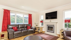 A living room with a leather sofa, ottoman, fireplace, TV, and large windows with curtains.