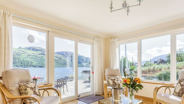 A room with large windows, wicker furniture, a glass table, and a view of a lake and hills.
