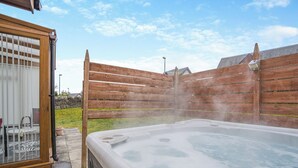 A hot tub in a backyard with a wooden fence, a glass door, and a stone wall in the background.