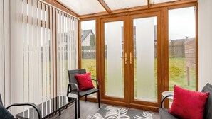 A sunroom with a view of a backyard, featuring a wooden-framed glass door, a black chair with a red pillow, and a grey sofa with a red pillow.