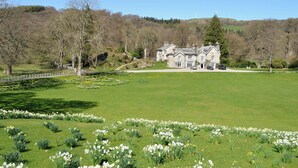 Un grand bâtiment en pierre, doté d’une façade symétrique, entouré d’une pelouse bien entretenue et agrémenté de diverses plantes à fleurs.