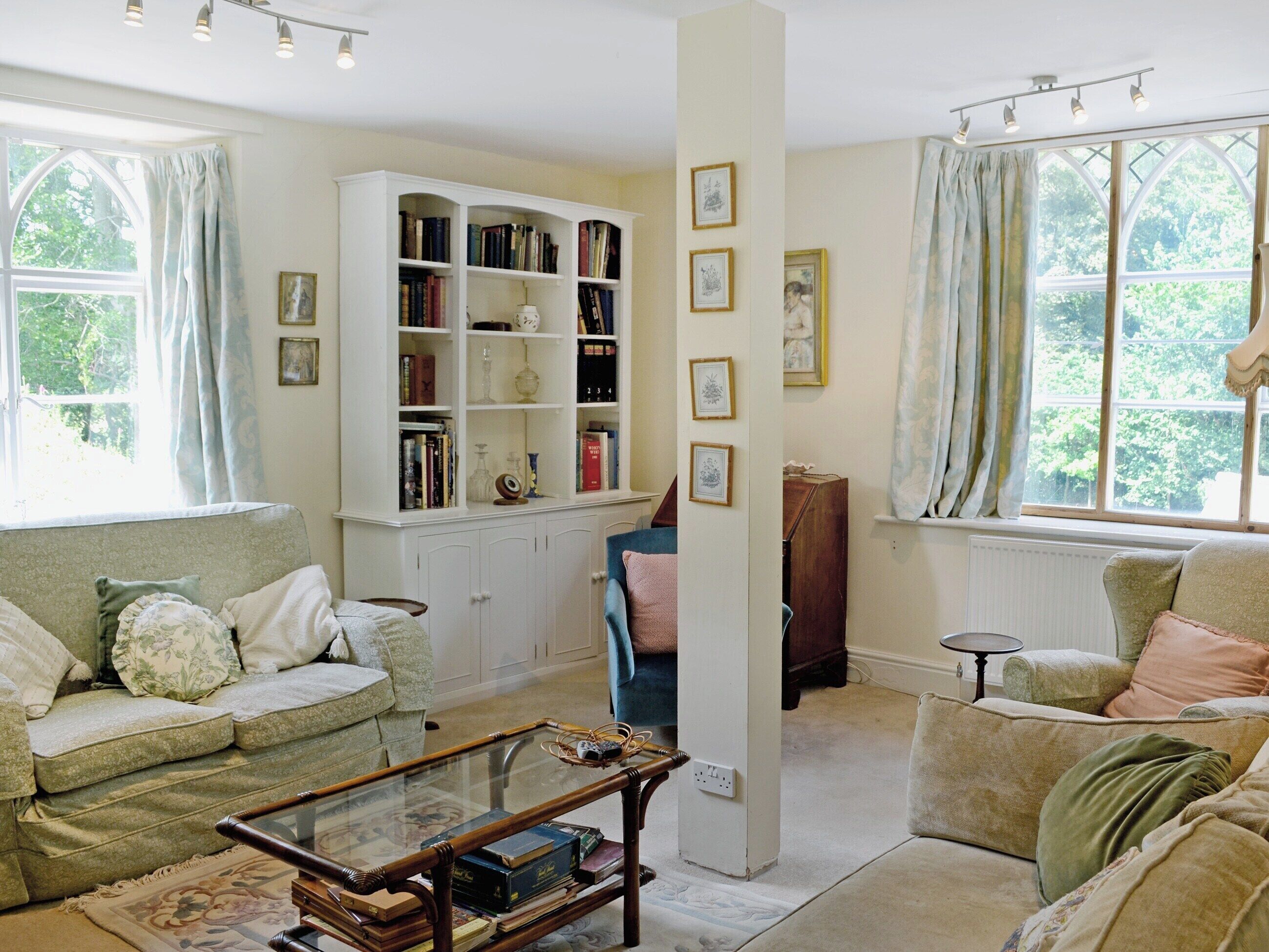 A cozy living room with a large white bookcase, two sofas, a glass coffee table, and a view of greenery through the windows.