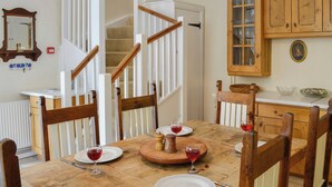 Un coin repas avec une table en bois dressée pour quatre personnes, entourée de chaises en bois. Un escalier avec des rampes blanches est visible au fond.