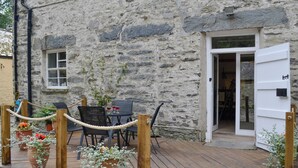 Un bâtiment en pierre doté d’une terrasse en bois, de sièges extérieurs et de plantes en pot.