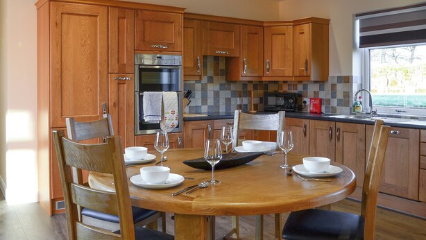 A modern kitchen with wooden cabinets, a built-in oven, and a dining area with a round table and chairs. - Liftingstane Cottage (Closeburn, near Thornhill)