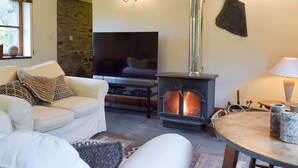 Un salon chaleureux avec un mur en pierre, un canapé blanc, un téléviseur, un poêle à bois et une lampe.