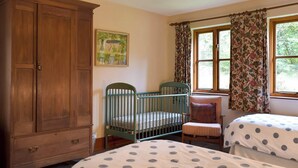 Une chambre avec deux lits simples, une armoire en bois, un berceau, une chaise et une fenêtre agrémentée de rideaux à motifs floraux.